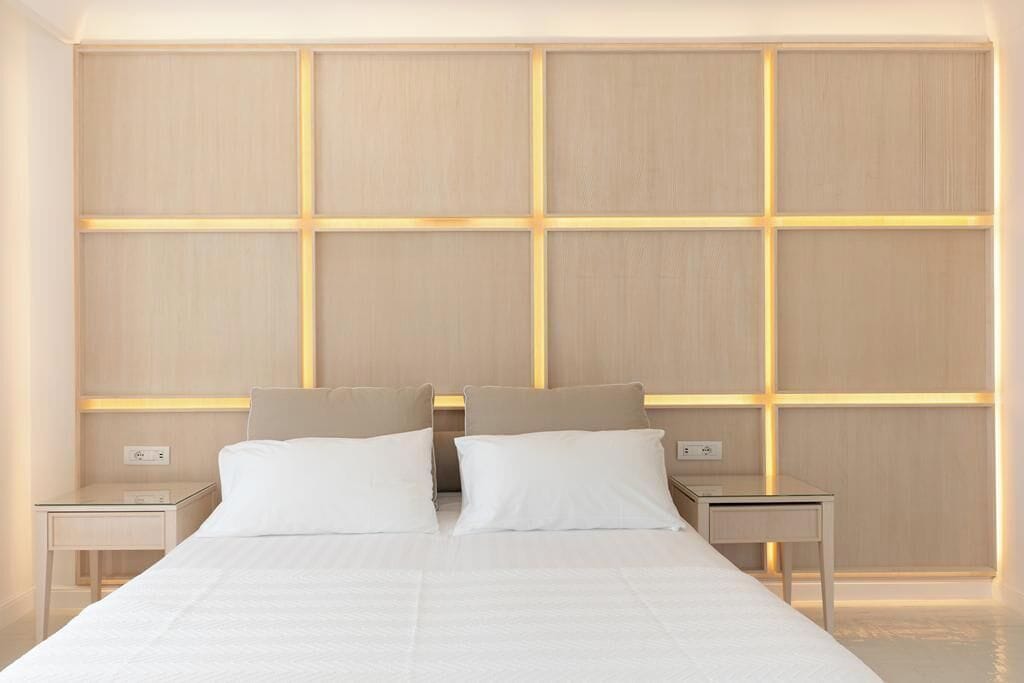 Una habitación de hotel moderna con una cama grande, dos mesitas de noche y un panel de madera con líneas iluminadas.