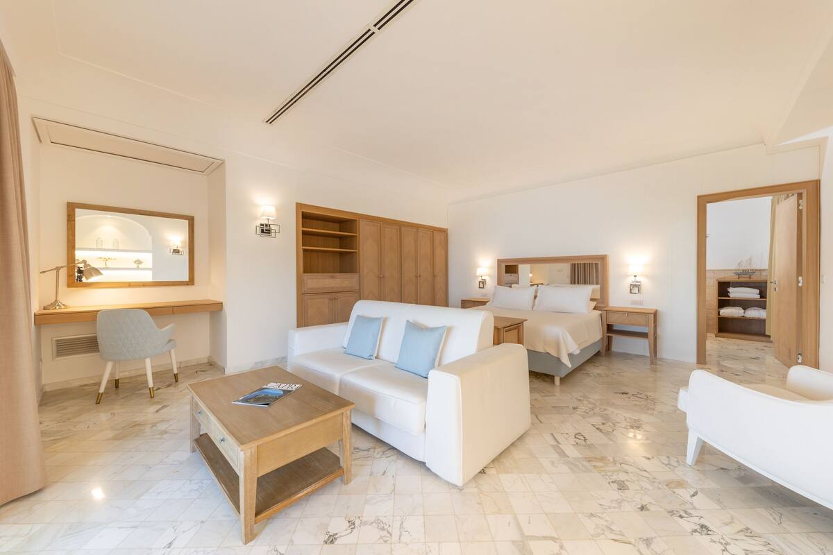 Una habitación de hotel moderna con un sofá blanco, una mesa de centro de madera, un escritorio con una silla, una cama con cabecera y un baño con bañera.