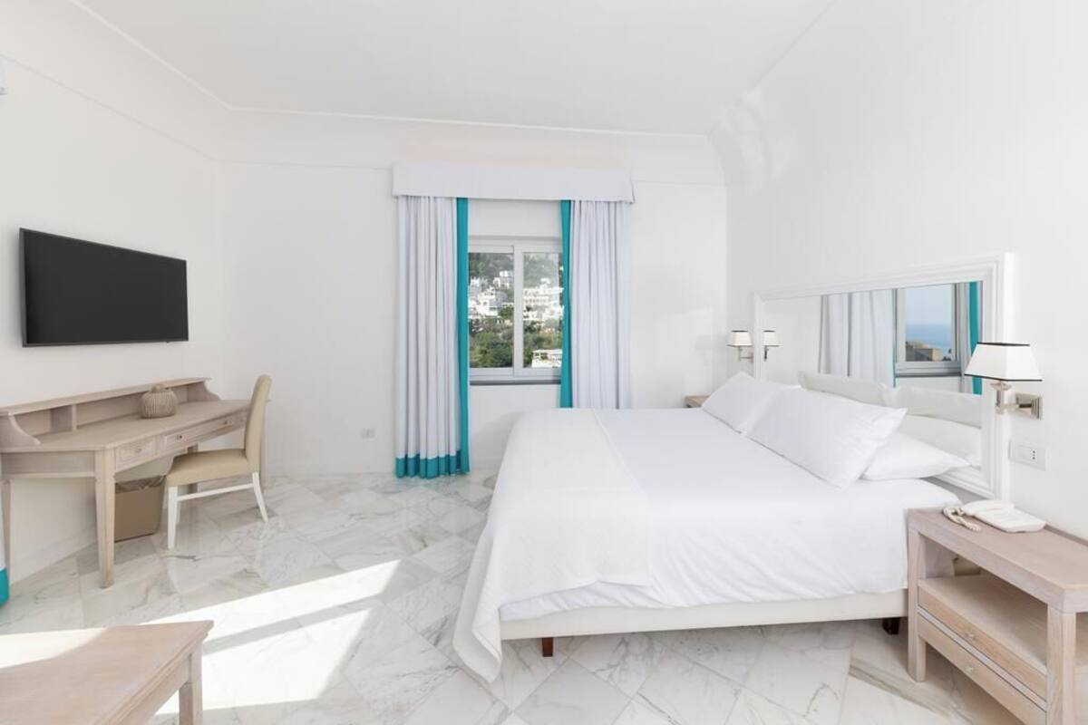 Habitación de hotel con cama, escritorio, silla, televisión y ventana con cortinas.