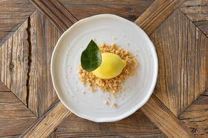 A white plate with a yellow dessert, a green leaf, and crumbled topping on a wooden table.