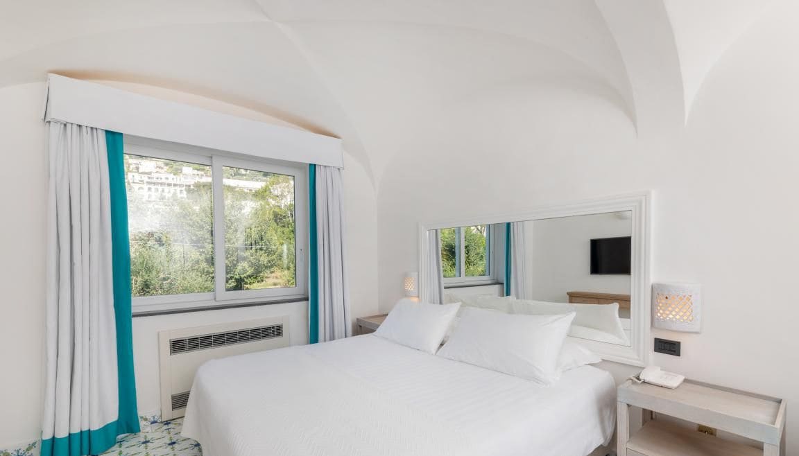 A hotel room with a large bed, a bedside table with a lamp, a window with curtains, and a wall-mounted TV.