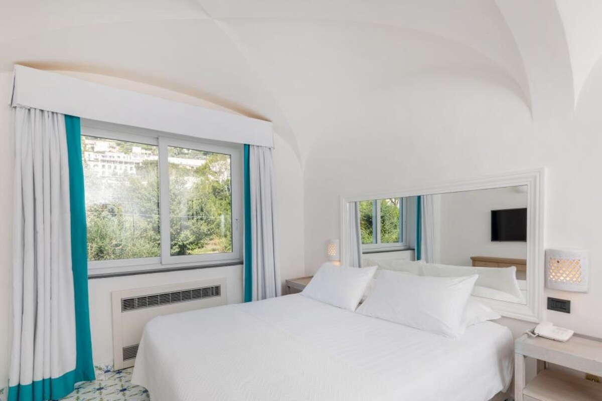 Una habitación de hotel con una cama grande, una mesita de noche con lámpara, una ventana con cortinas y un televisor montado en la pared.