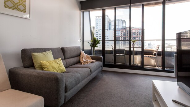 A modern living room with a grey sofa, a small table, and a large window offering a city view. - ABC Accommodation - 534 Flinders (Melbourne)