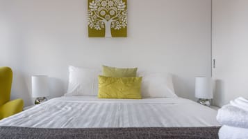 A hotel room with a bed, two bedside lamps, a yellow armchair, a green pillow, and a wall art piece.