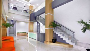 A modern hotel lobby with a reception desk, bright orange seating, a staircase, and potted plants.