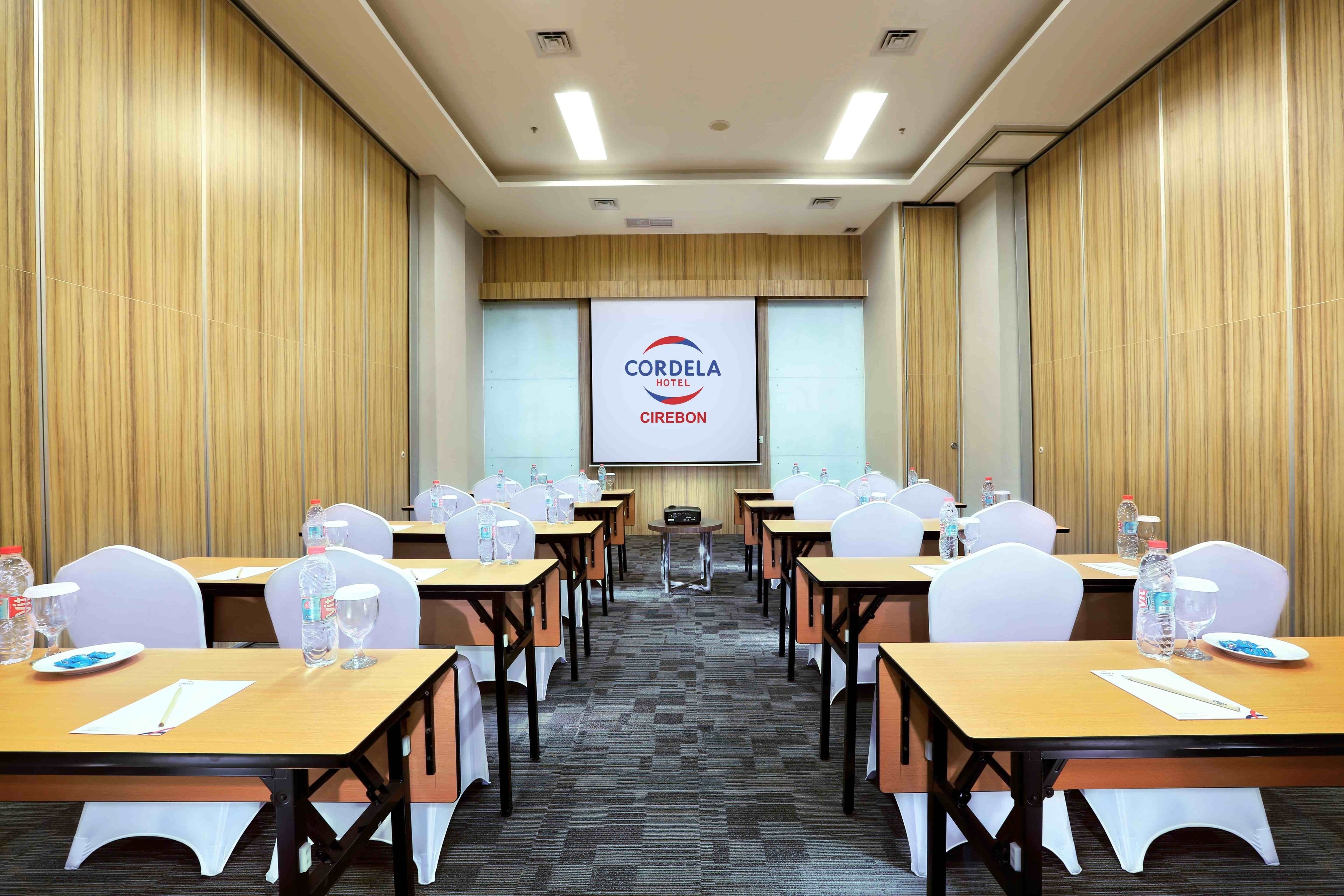 A conference room with tables, chairs, and a projection screen displaying 'CORDELA HOTEL CIREBON'.