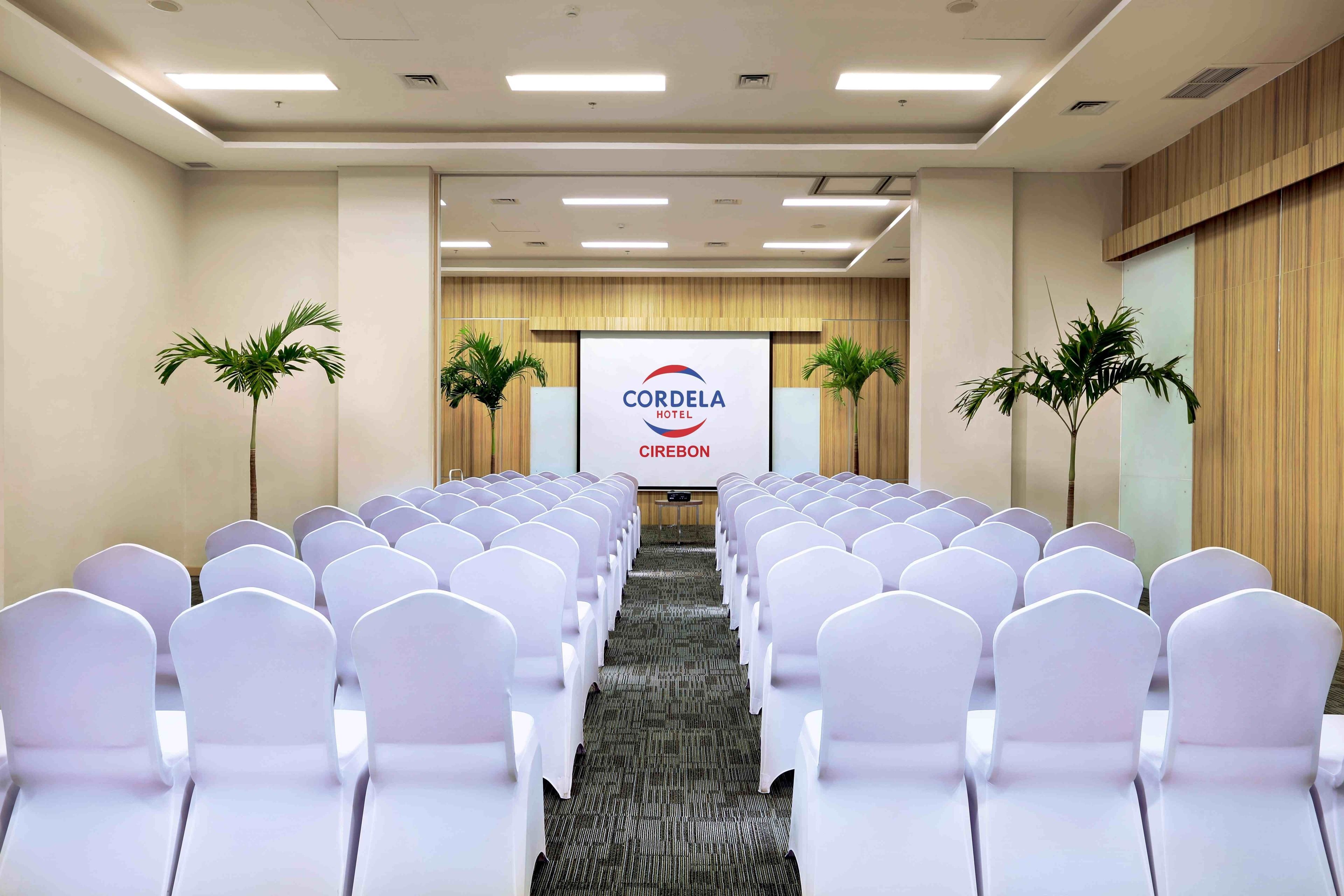 A conference room with rows of white chairs, a podium, and a screen displaying 'CORDELA HOTEL CIREBON'.