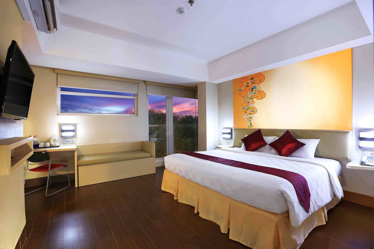 Ein Hotelzimmer mit einem großen Bett, einem Schreibtisch mit Stuhl, einem Fernseher und einem Fenster mit Blick auf die Stadt.