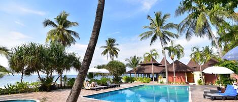 A pool area with lounge chairs and umbrellas, surrounded by palm trees and a view of the sea.