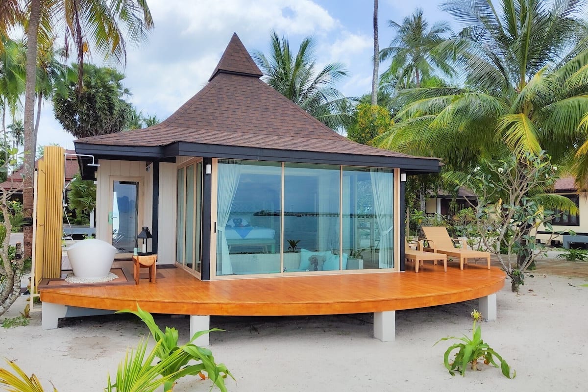 Een modern strandhuis met een kamer met glazen wanden, een houten terras en palmbomen.