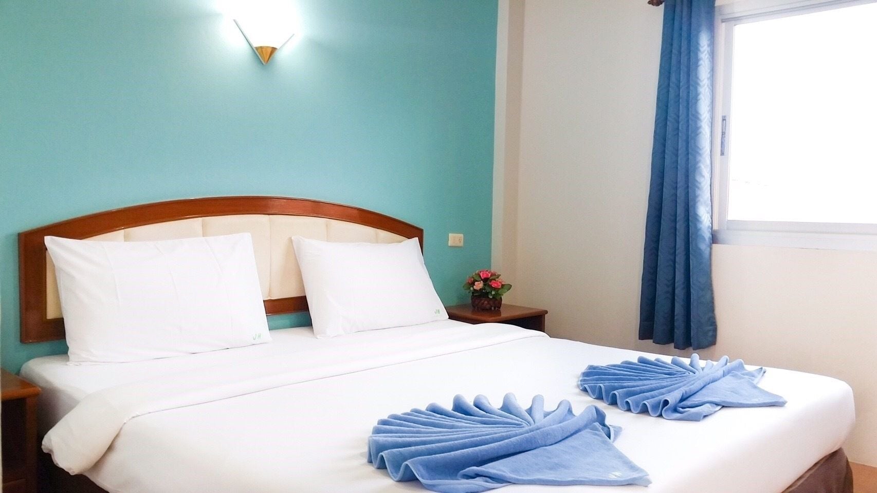 Una cama bien hecha con sábanas blancas y toallas azules, un cabecero de madera, una mesita de noche con un florero y una ventana con cortinas azules.
