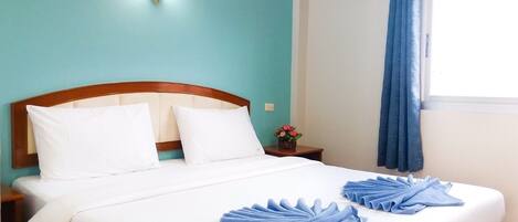 Una cama bien hecha con sábanas blancas y toallas azules, un cabecero de madera, una mesita de noche con un florero y una ventana con cortinas azules.