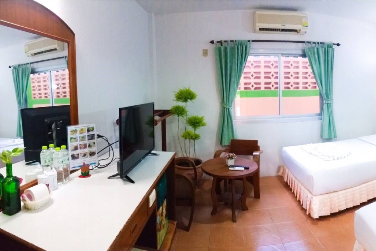 A hotel room with a bed, a desk with a computer, a chair, a small table with a plant, and a window with curtains.