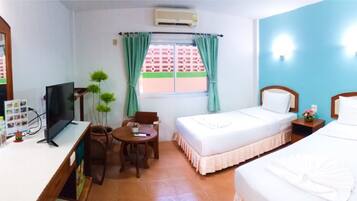 A hotel room with a bed, a desk with a computer, a chair, a small table with a plant, and a window with curtains.