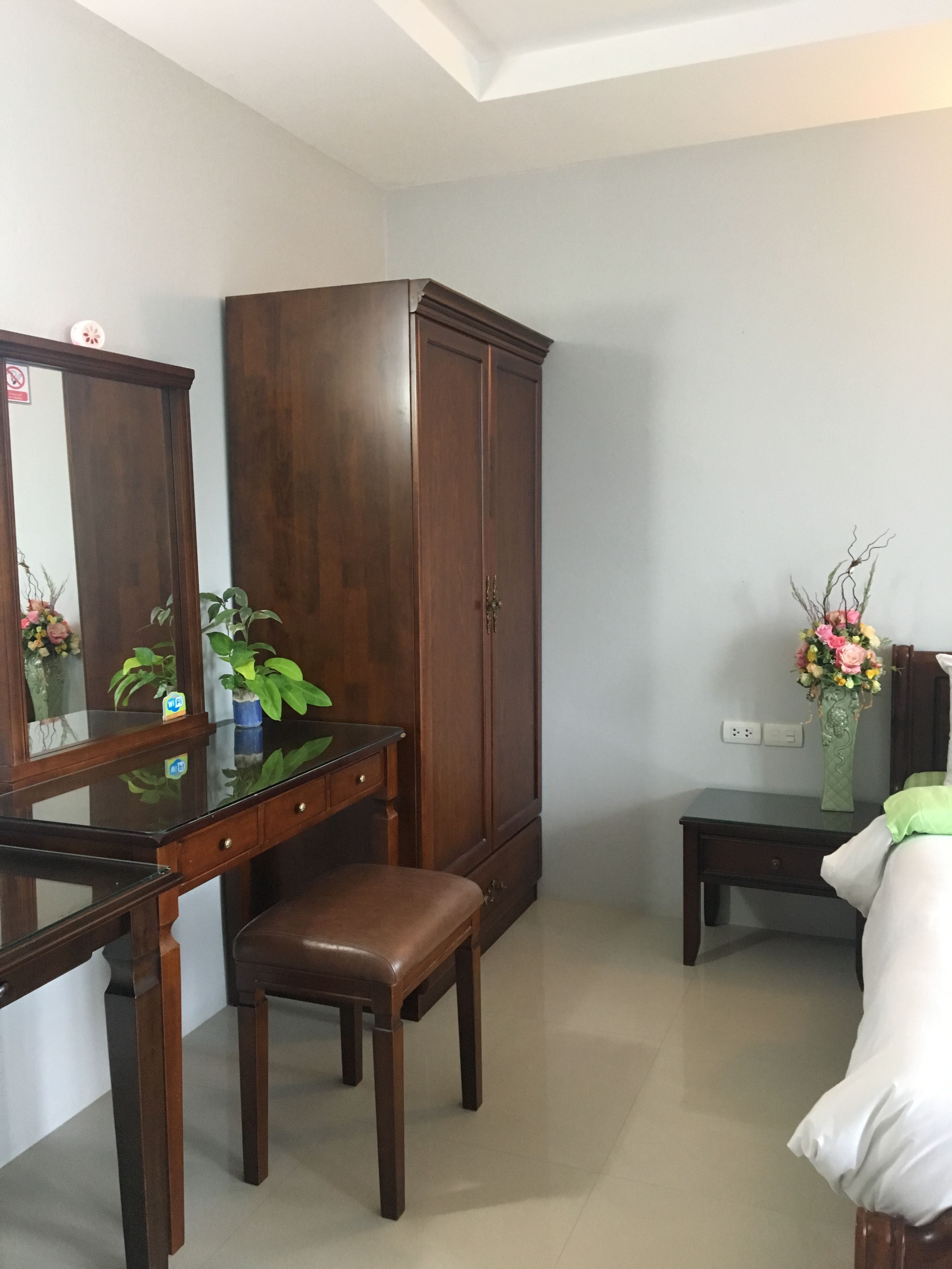 A room with a wooden desk, a chair, a wardrobe, a bedside table with a vase of flowers, and a bed.