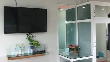 Une pièce dotée d’un téléviseur à écran plat fixé au mur, d’une petite étagère avec des bouteilles d’eau et une plante, d’une vitrine contenant des fleurs et d’une poubelle.