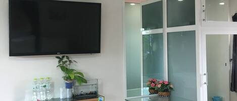 A room with a flat-screen TV mounted on the wall, a small shelf with water bottles and a plant, a glass cabinet with flowers, and a trash bin.