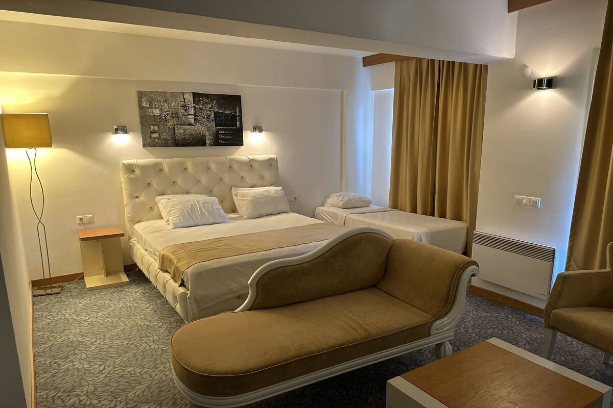 A hotel room with a bed, a sofa, a small table, a lamp, and a wall-mounted heater.