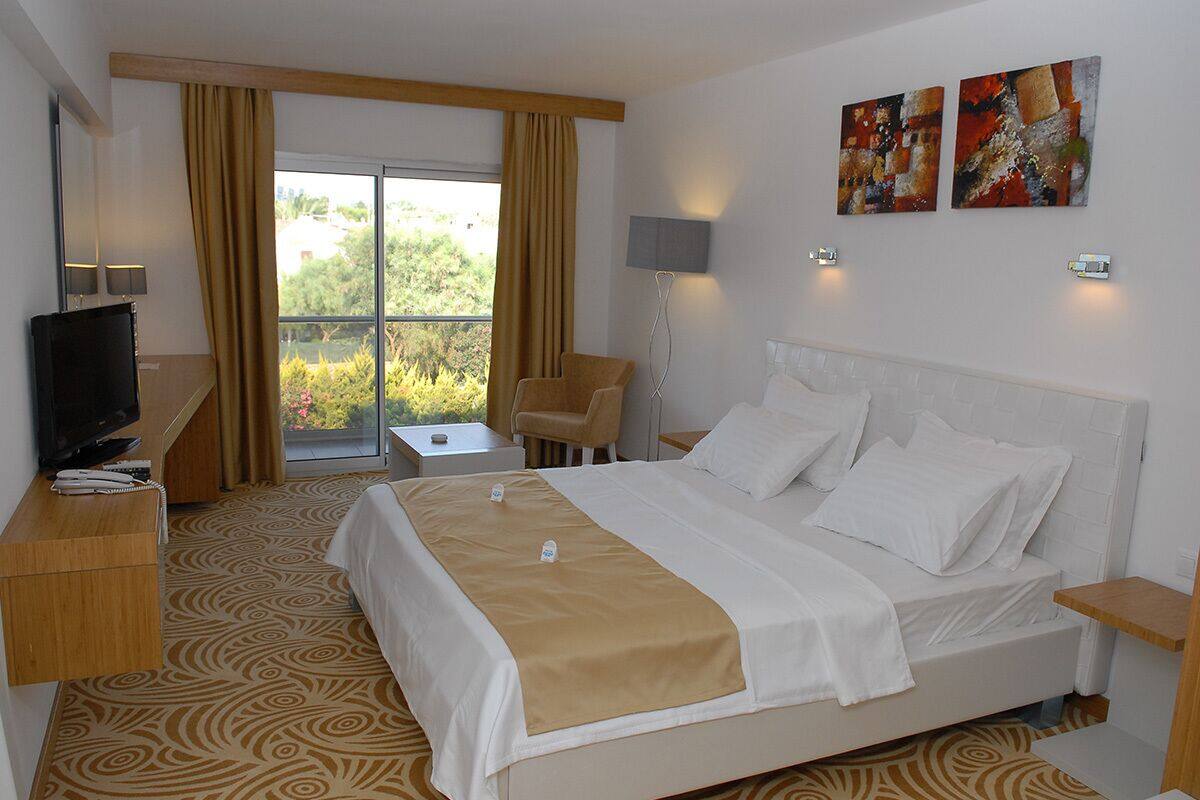 A hotel room with a large bed, a desk with a TV, a chair, and a balcony with curtains.
