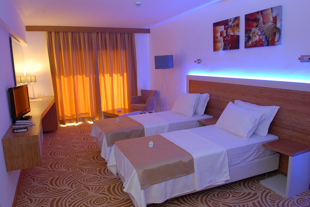 Ein Hotelzimmer mit zwei Einzelbetten, einem Schreibtisch mit Lampe, einem Stuhl und einem großen Fenster mit Vorhängen.