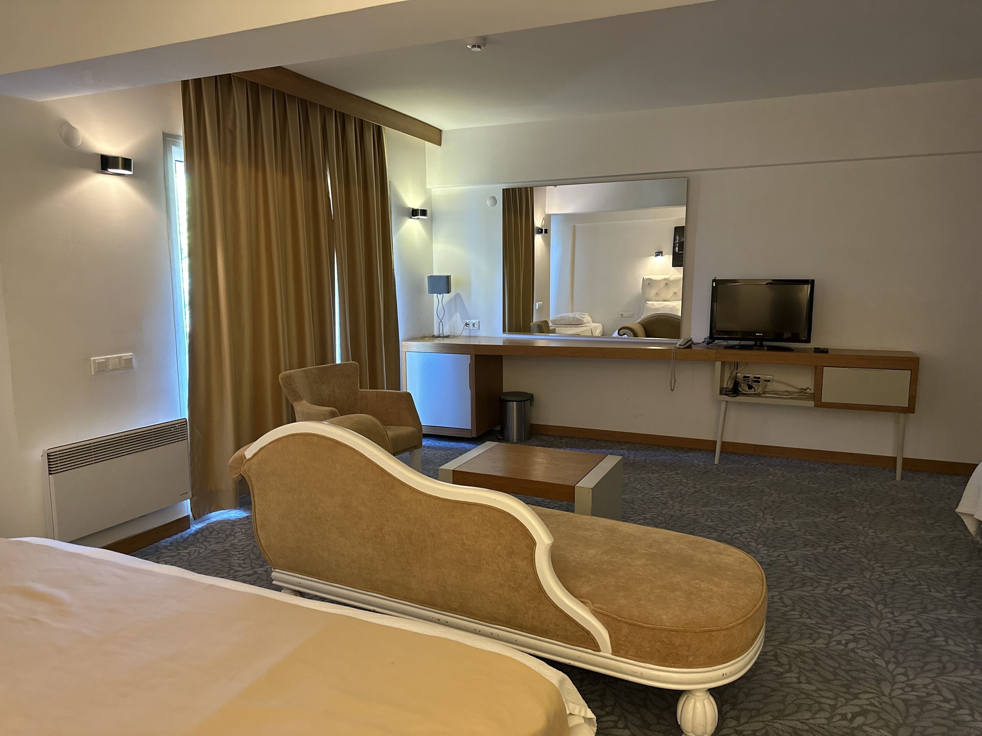 Habitación de hotel con cama, zona de estar con una silla, escritorio con televisor y espejo.
