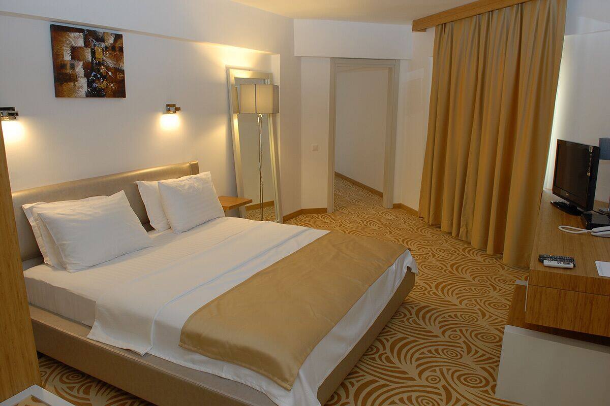 A hotel room with a bed, bedside table, lamp, TV, and a large window with curtains.