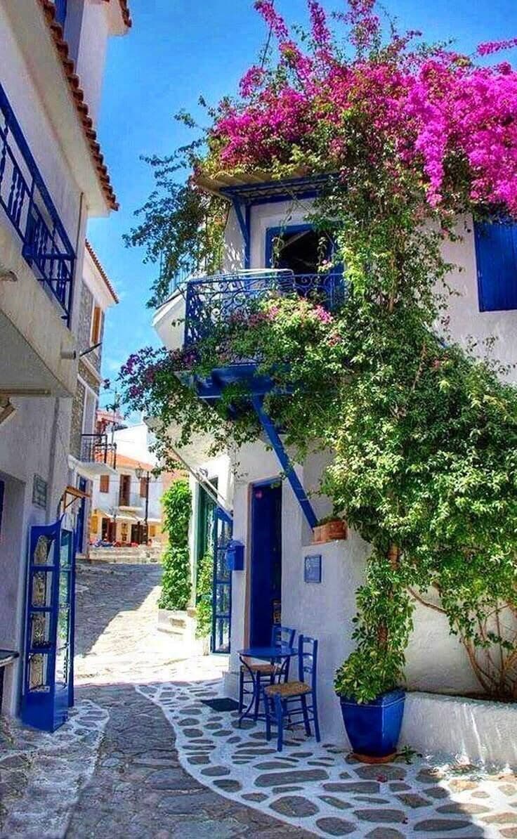 A narrow street with a building featuring a balcony with pink flowers, a blue door, and a stone-paved path.