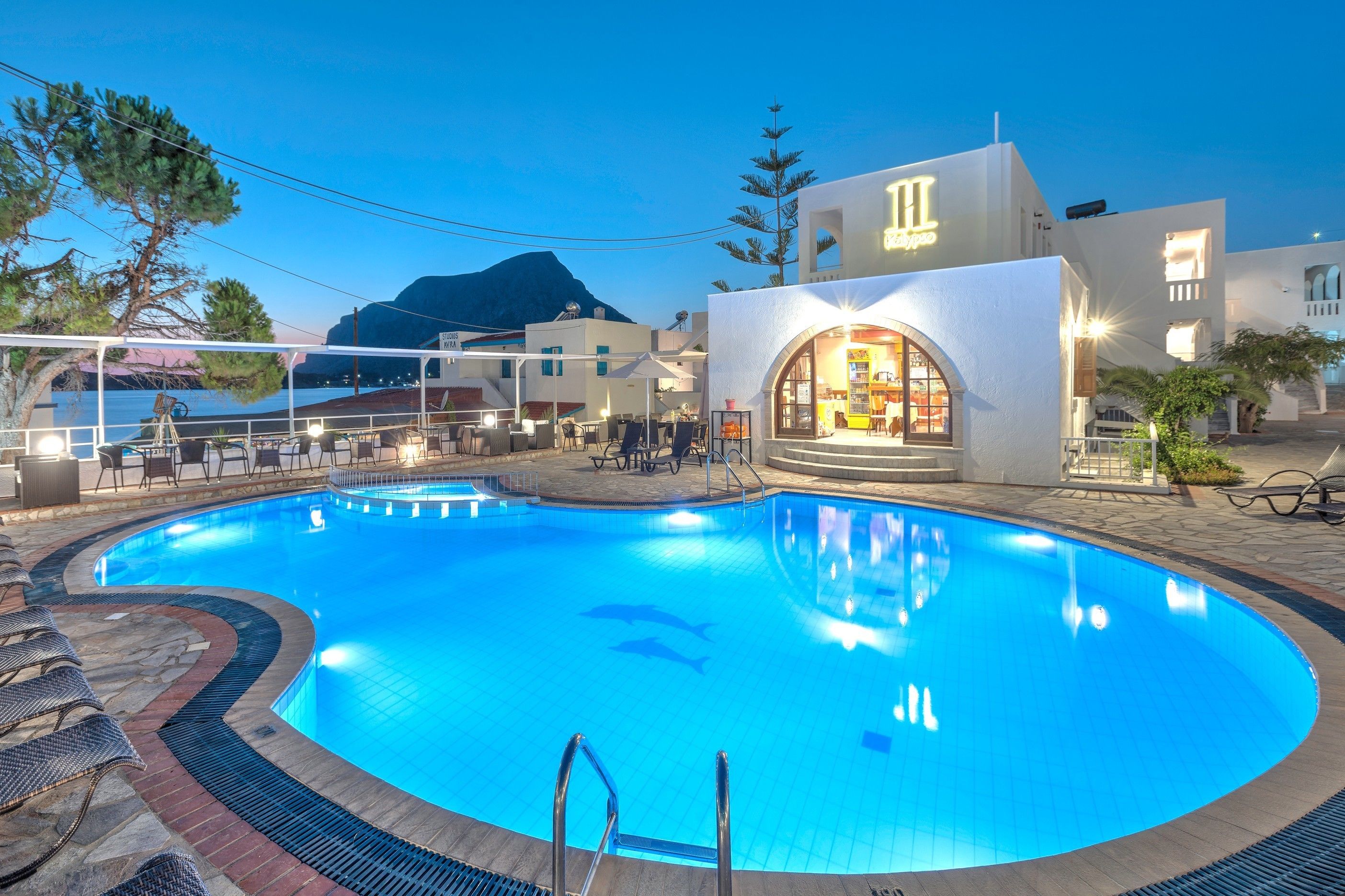Una piscina con sillones de descanso, un restaurante y una montaña al fondo.