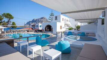 Un área de piscina con sillones de descanso y un jacuzzi, rodeada de edificios blancos y vistas a la montaña.