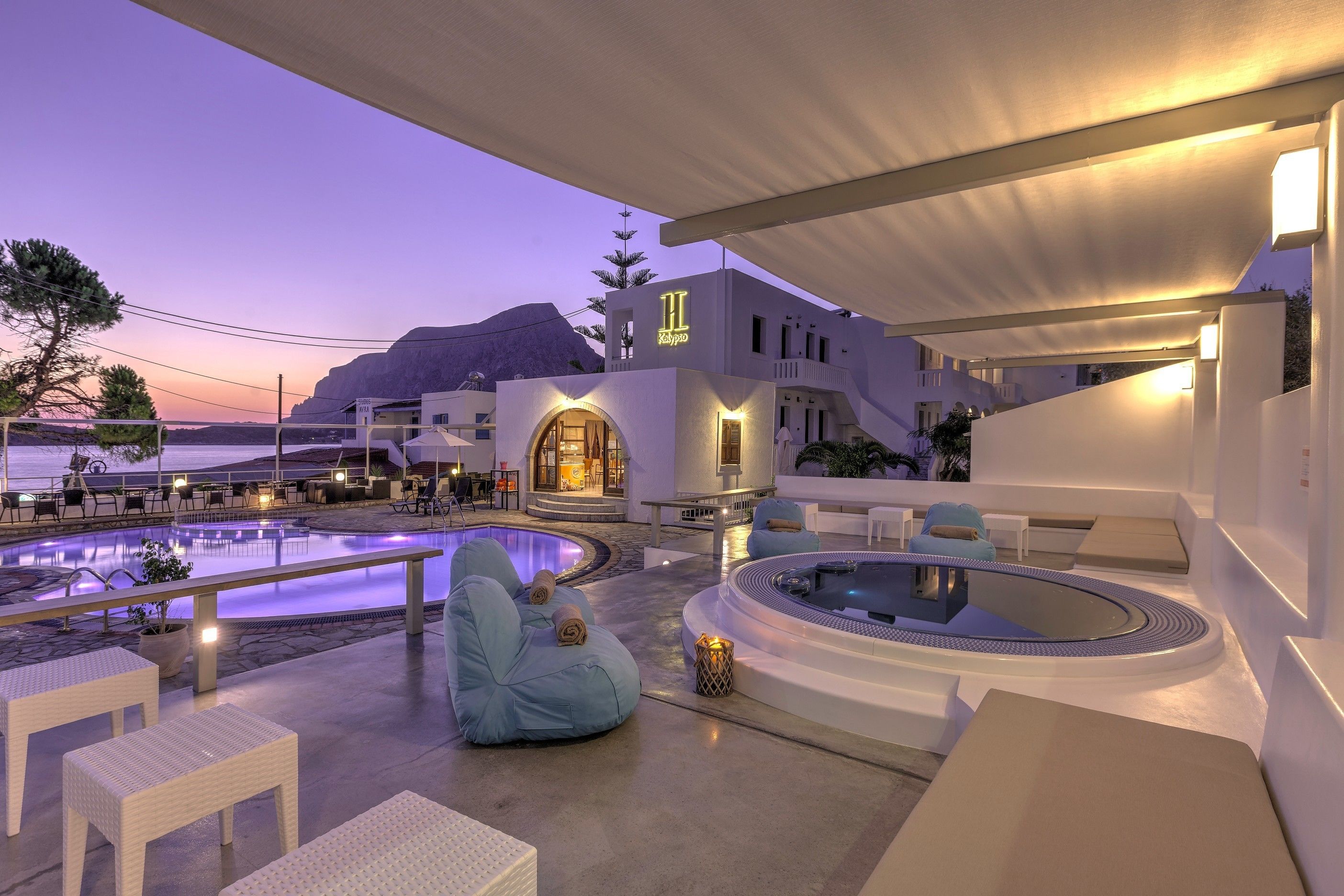 A modern outdoor pool area with seating and a hot tub, surrounded by white buildings with a mountain in the background.