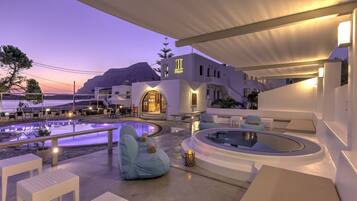 A modern outdoor pool area with seating and a hot tub, surrounded by white buildings with a mountain in the background.