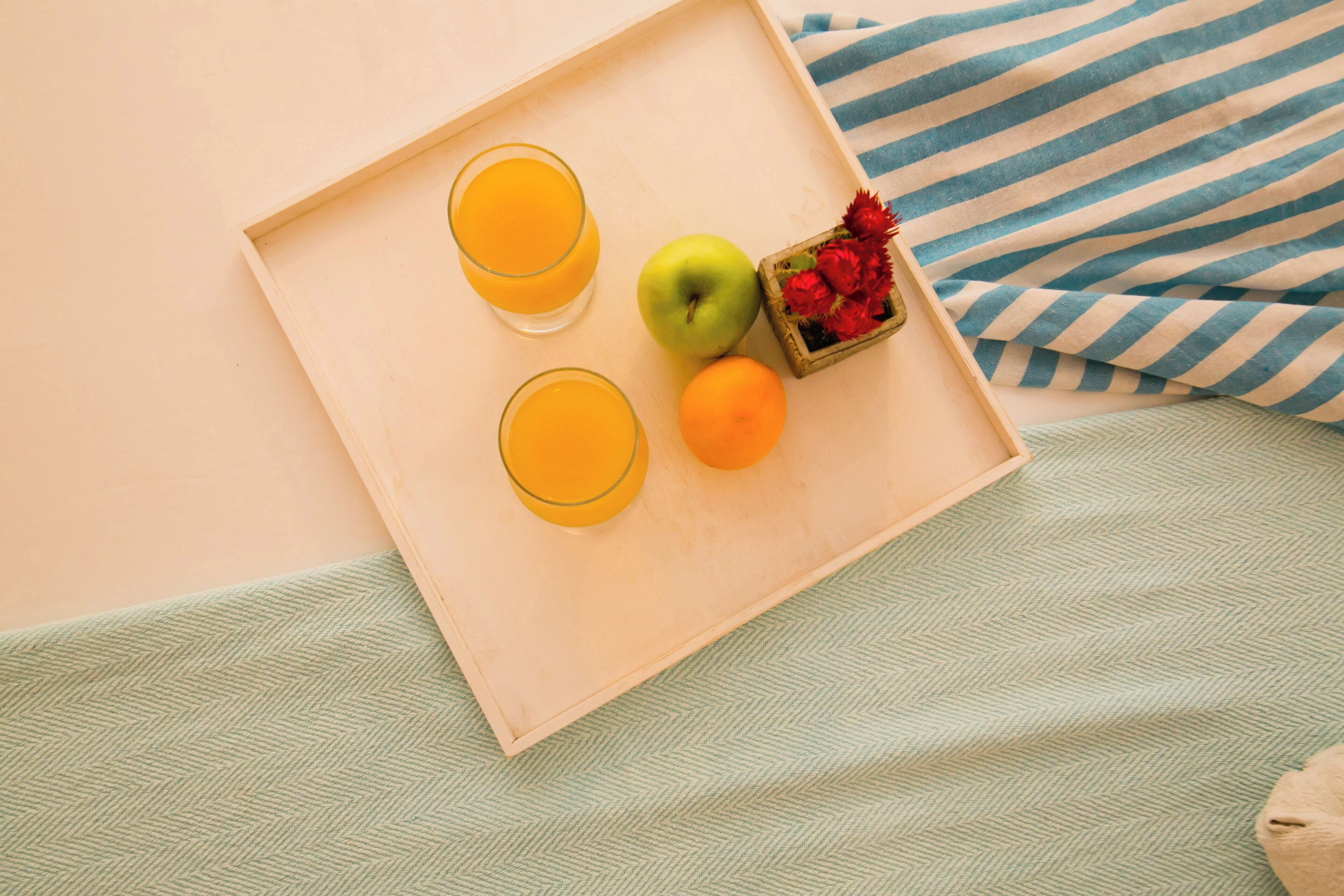 Un plateau en bois avec deux verres de jus d’orange, une pomme et une orange, posés sur un lit recouvert d’une couverture verte à texture et agrémenté d’oreillers à rayures.