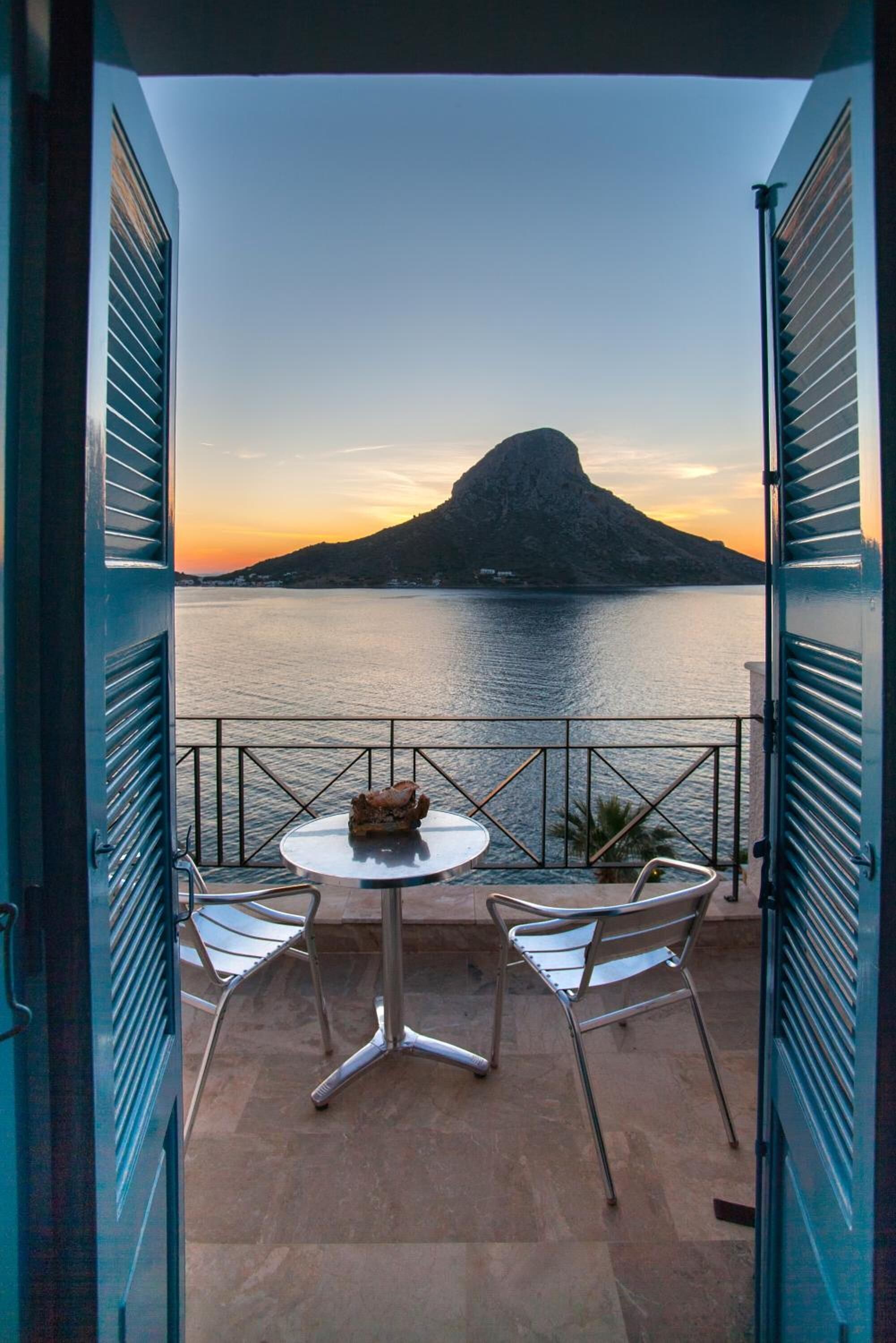 An open balcony with a table and two chairs, overlooking a body of water and a mountain at sunset.