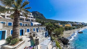 Un edificio costero con puertas y ventanas azules, una palmera y vistas al mar y una montaña.
