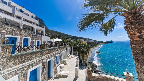 Un edificio costero con muros de piedra, puertas azules y mesas al aire libre frente al mar.