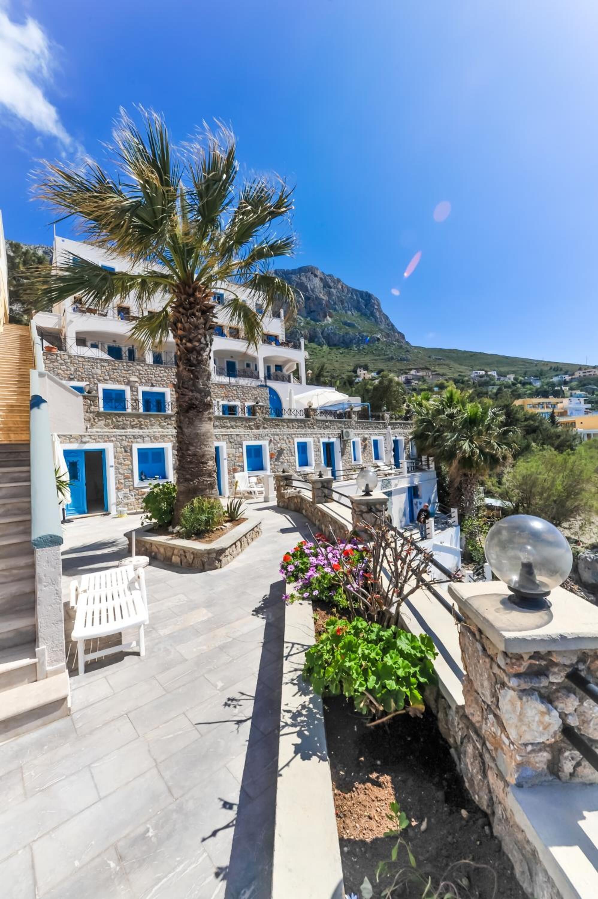 Un edificio costero con puertas y ventanas azules, una palmera y vistas al mar y las montañas.