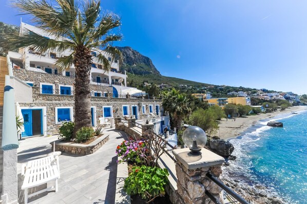 A coastal building with stone walls, blue shutters, and a palm tree. - Ambiance Studios (Kalymnos)