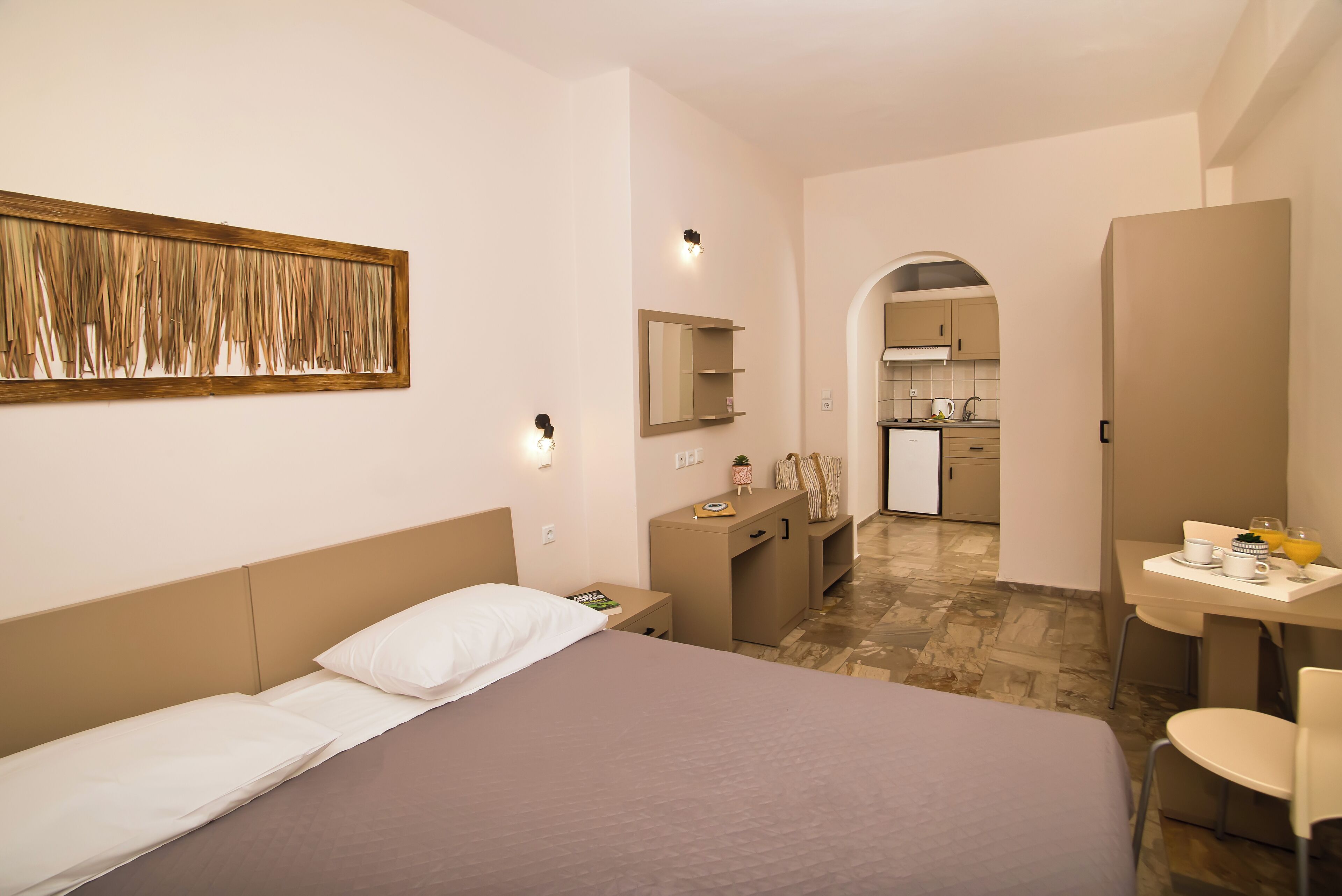 Una habitación de hotel moderna con una cama grande, un escritorio, una silla, una pequeña zona de cocina y un baño visible a través de un arco.