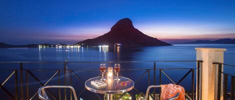 Une table avec deux chaises et deux verres de vin, donnant sur une étendue d’eau calme avec une montagne en arrière-plan.