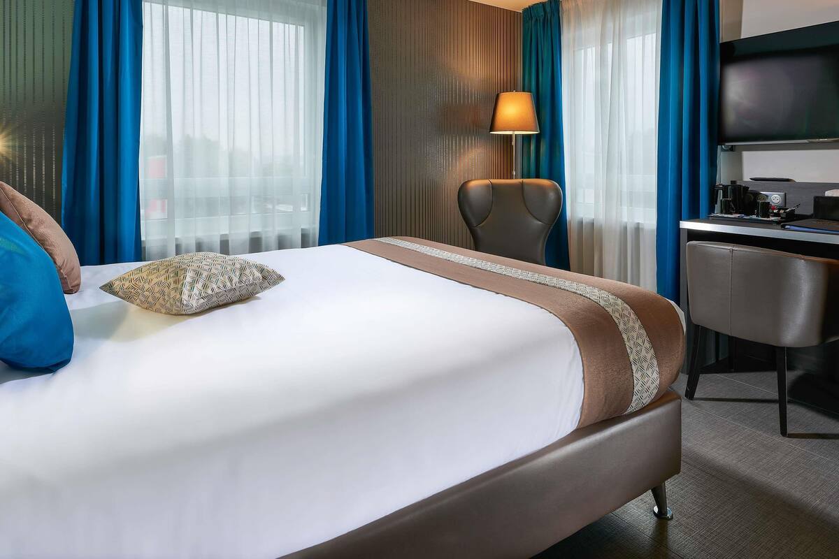 A hotel room with a bed, a desk, a chair, a TV, and curtains.
