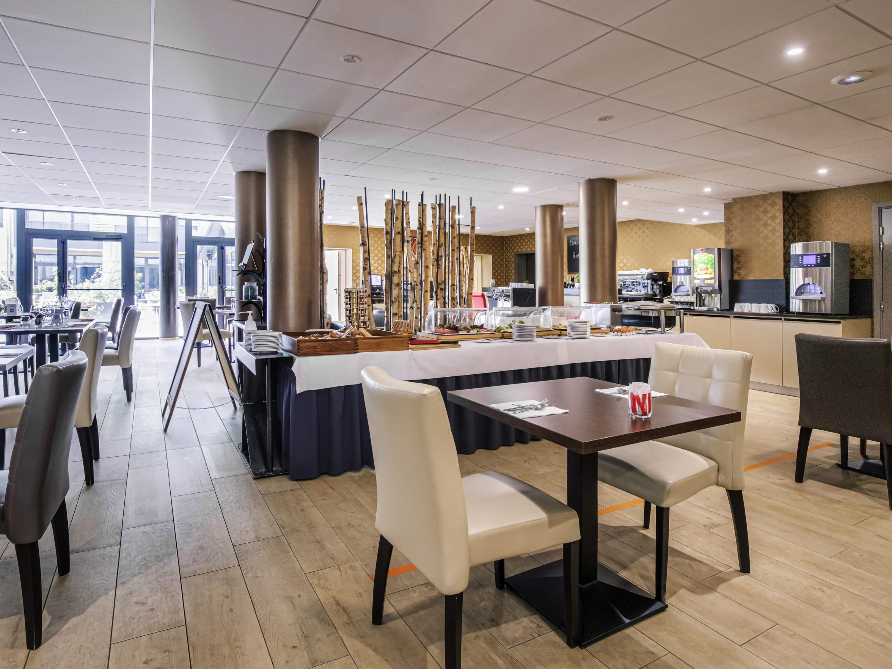 Ein Speisesaal eines Hotels mit Tischen, Stühlen, einer Buffetstation und einer Küche im Hintergrund.