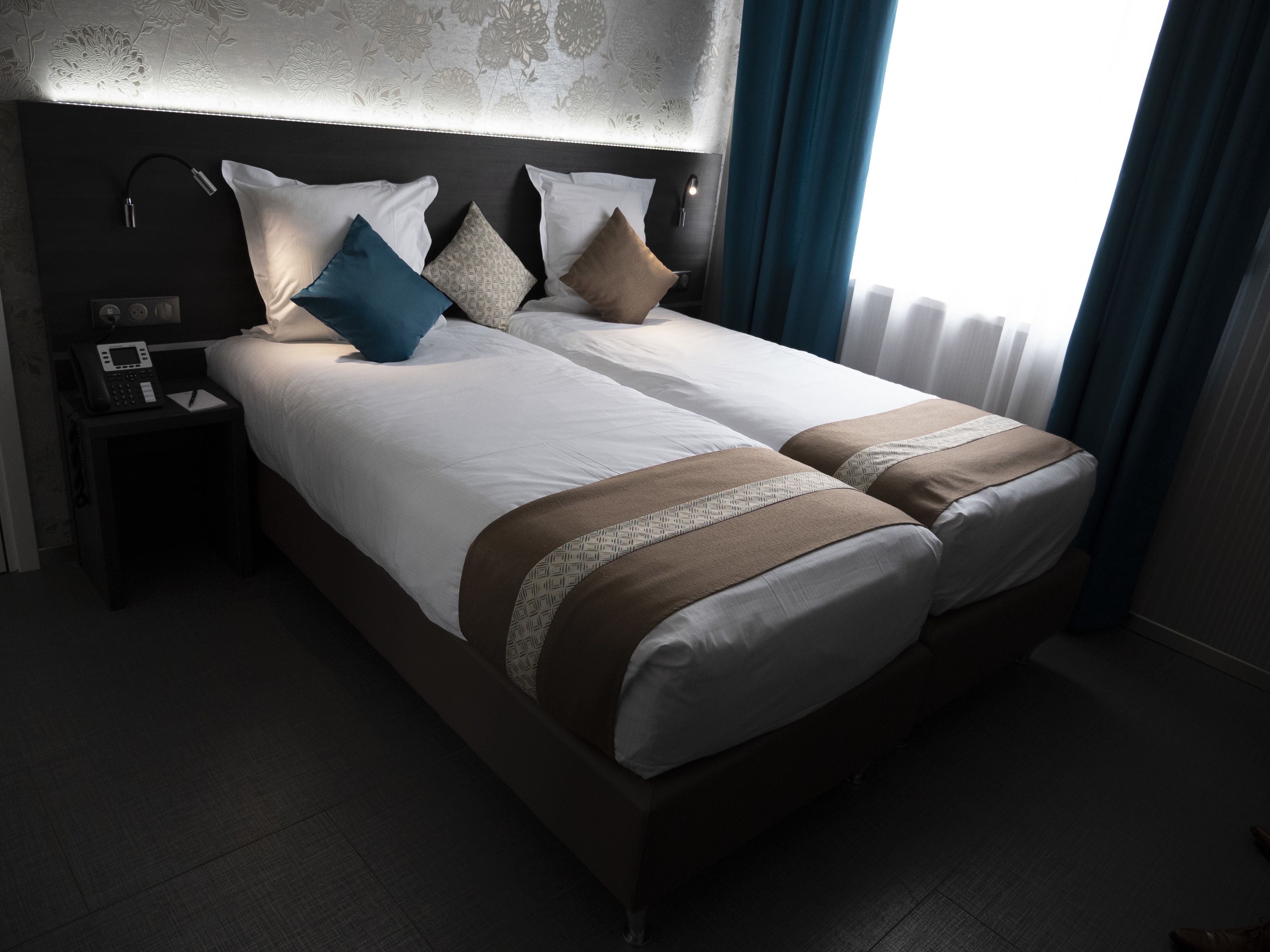 Cama doble con ropa de cama blanca y marrón, mesita de noche con teléfono y libreta, y cabecera con lámpara.