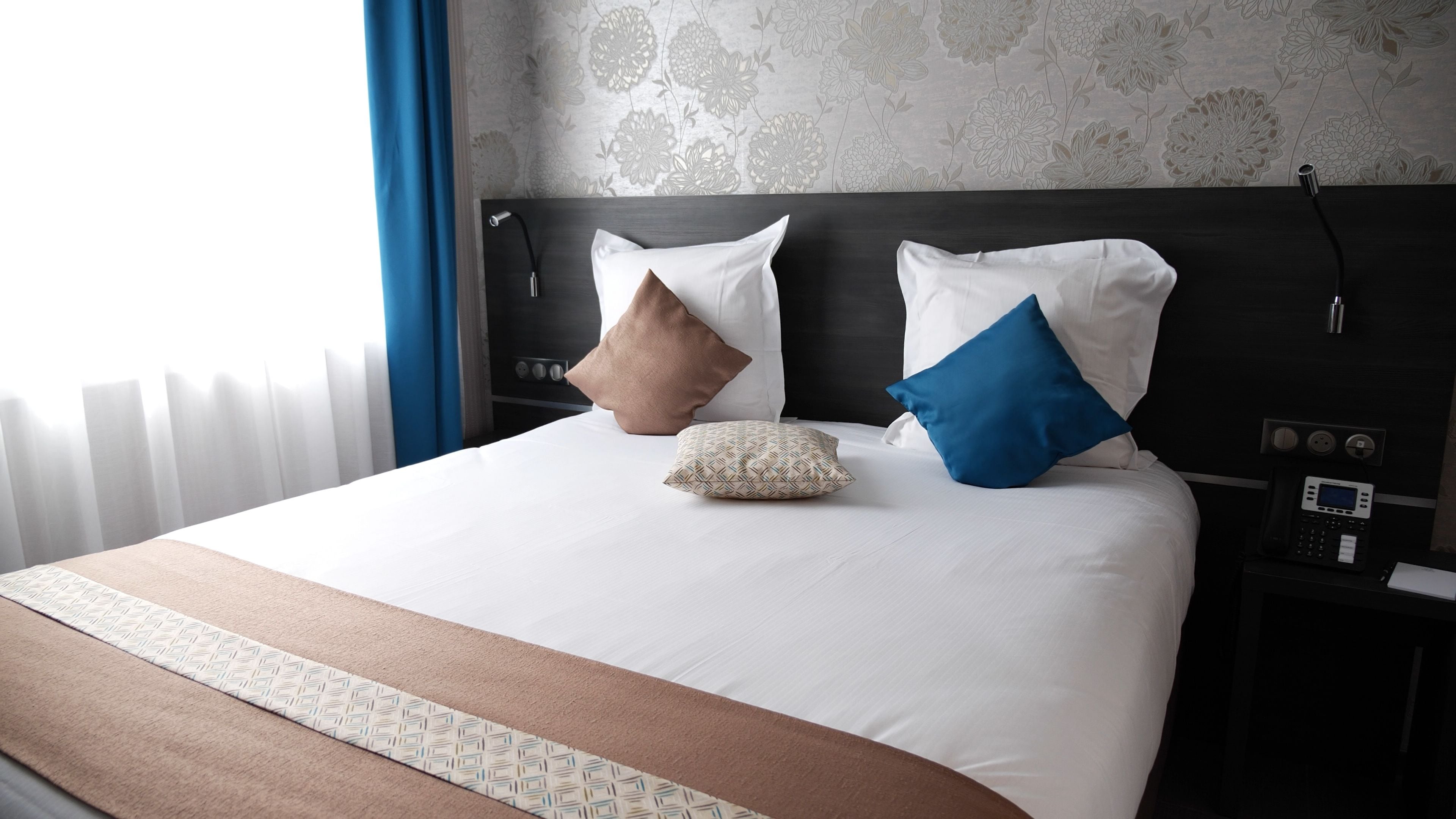 A hotel room with a large bed, two pillows, a bedside table with a phone, and a wall with floral wallpaper.
