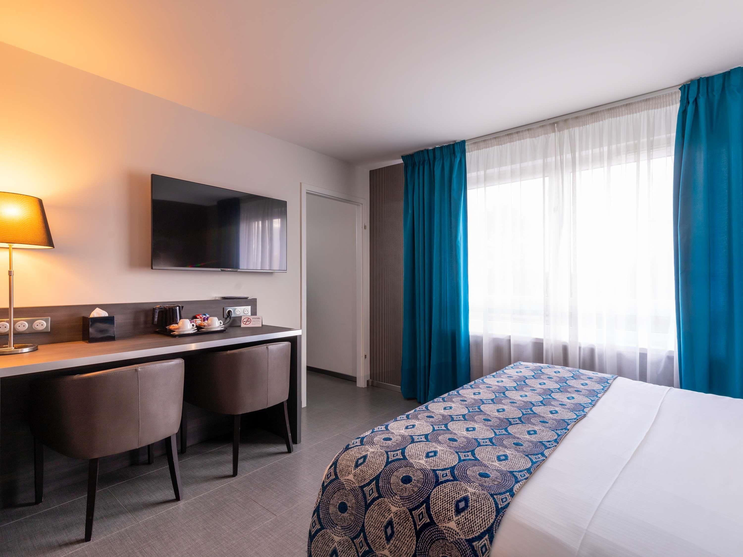 Ein Hotelzimmer mit Bett, Schreibtisch mit Stühlen, Fernseher und einem Fenster mit Vorhängen.