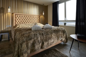 Un dormitorio con cabecera capitonada, cama cubierta de piel, mesita de noche y ventana con cortinas.