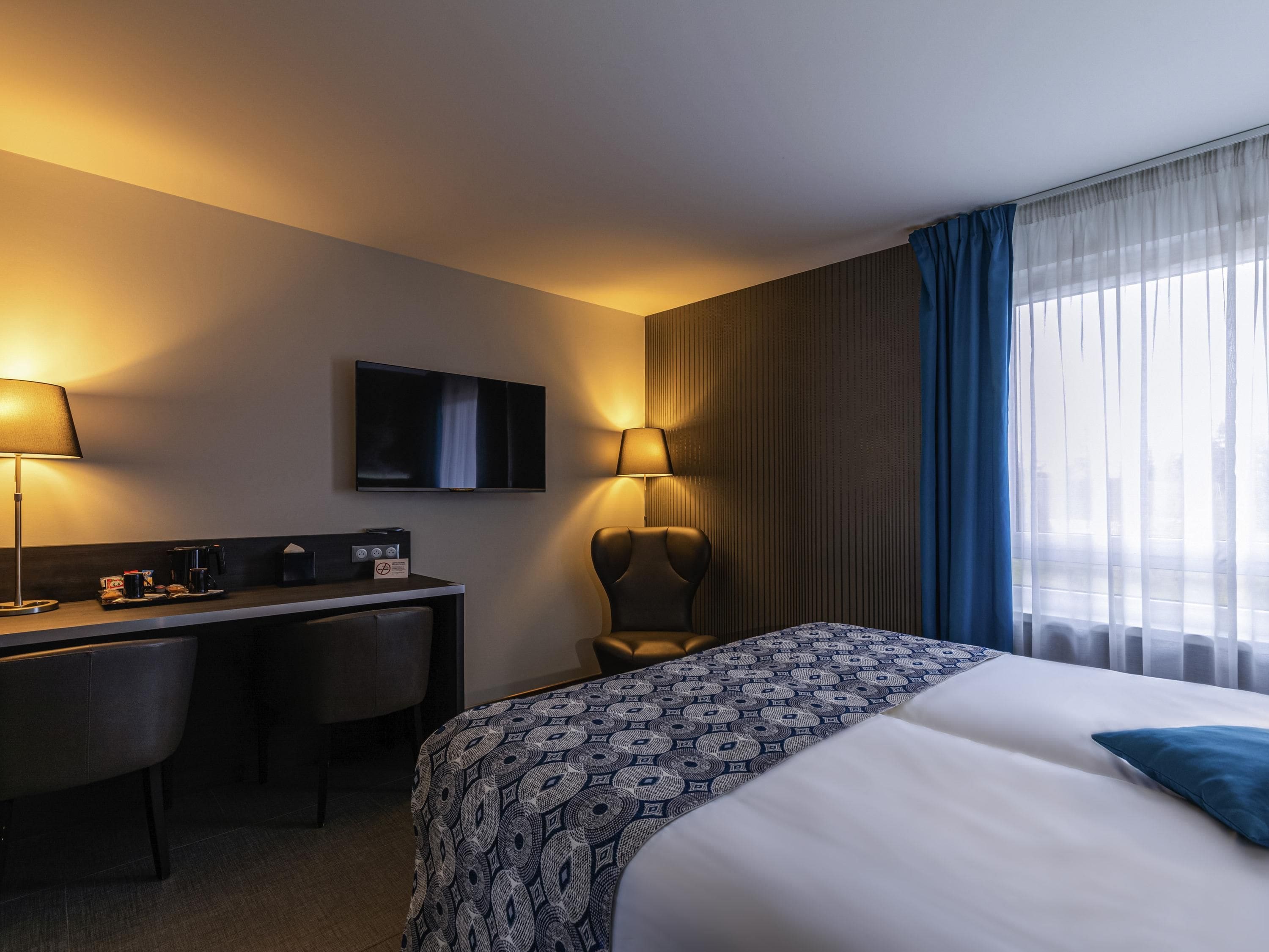 Una habitación de hotel con cama, escritorio, silla, televisor y cortinas.