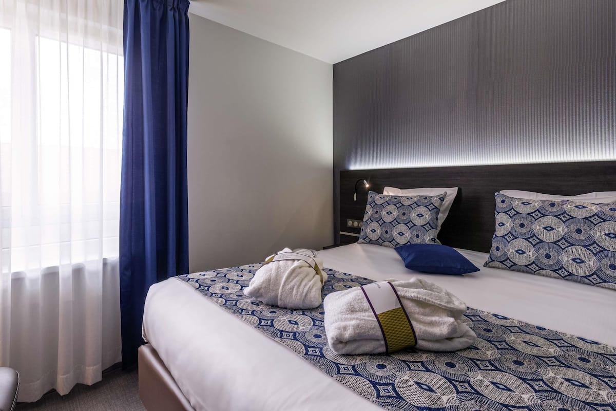 A hotel room with a large bed, a headboard with built-in lighting, a window with sheer curtains, and a dark wall.