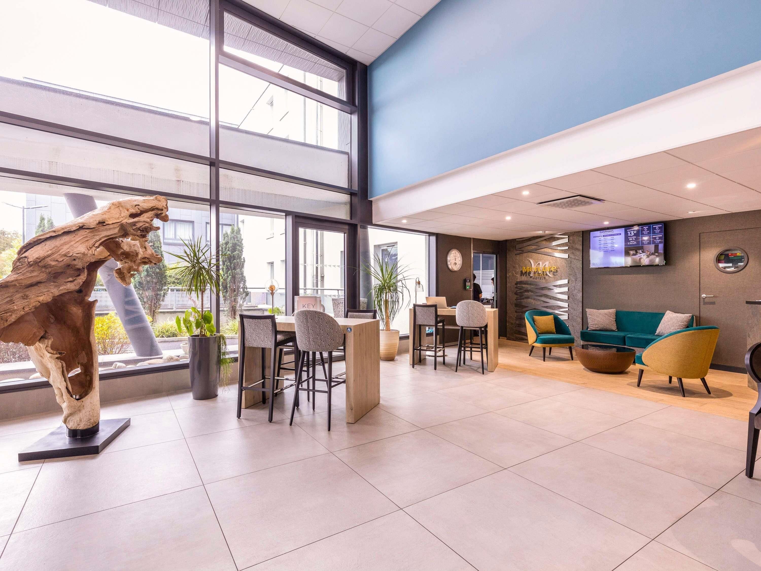 A modern hotel lobby with large windows, a reception desk, seating area, and a mounted TV.