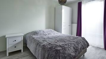 A bedroom with a bed, a white nightstand with drawers, a wardrobe, and a window with purple curtains.