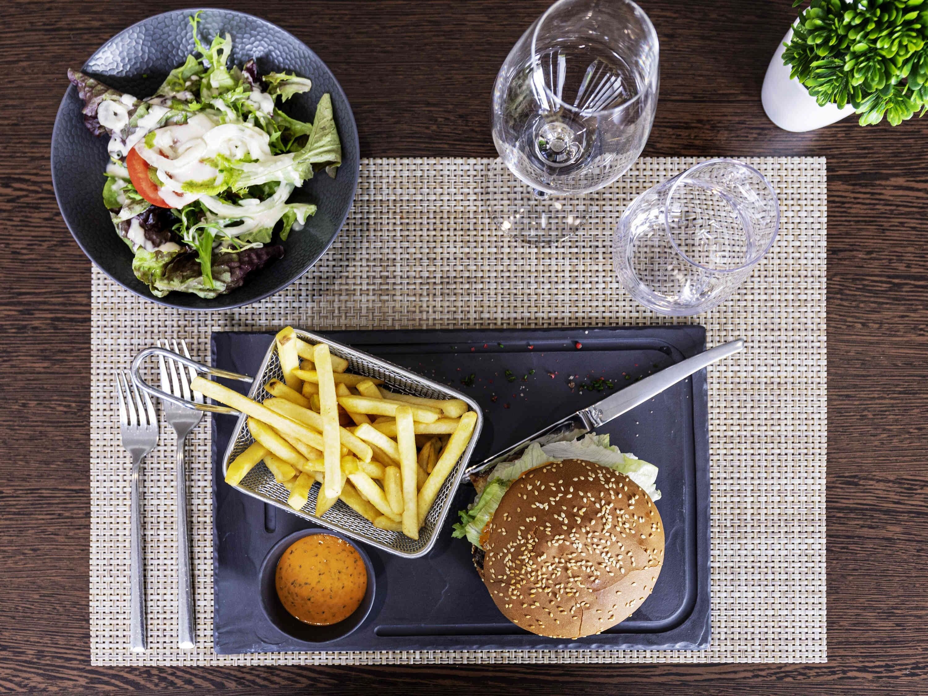 A meal tray with a burger, fries, and salad, a fork, knife, and a small bowl of sauce, on a table with a woven placemat.